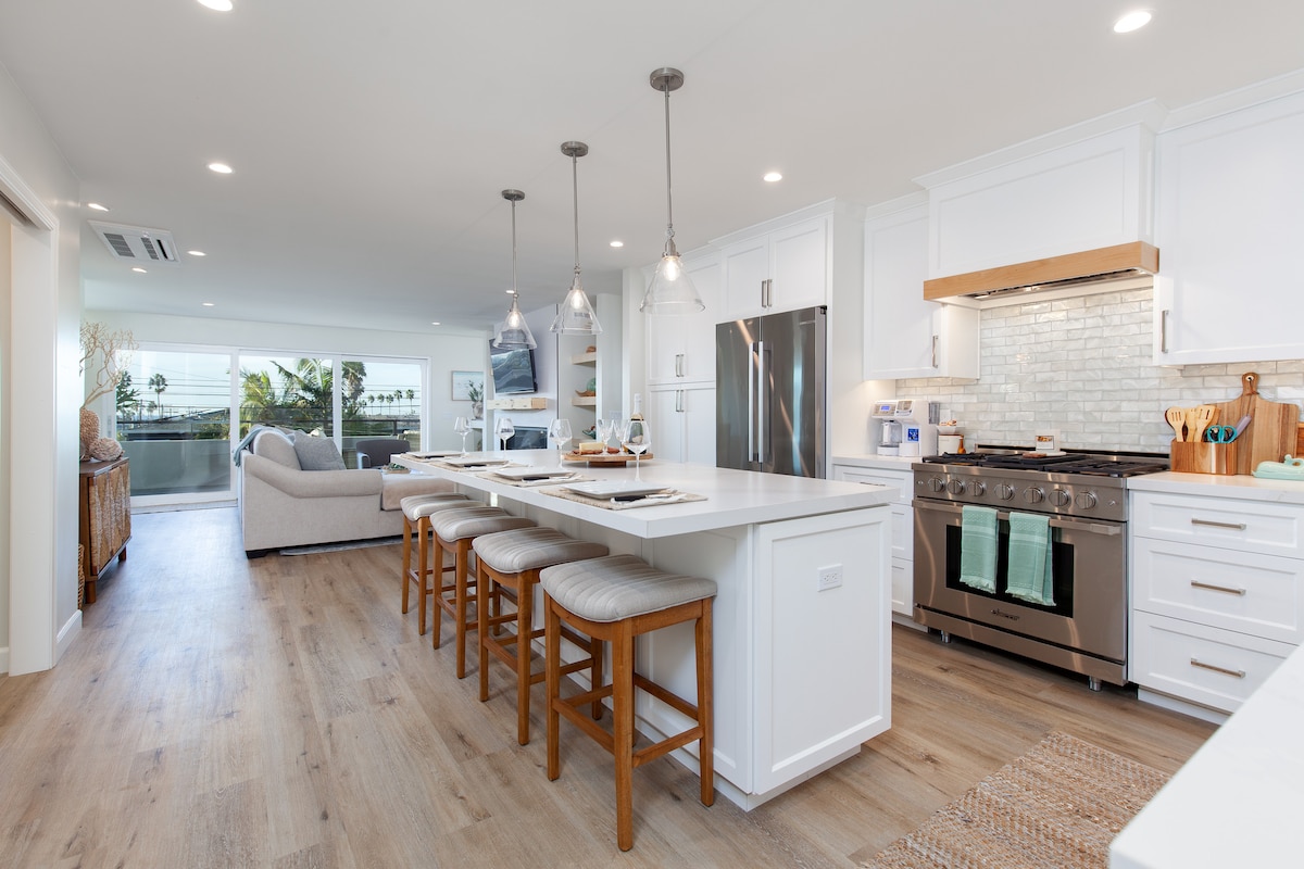 A modern kitchen features an expansive island with four high-backed stools and hanging pendant lights. Stainless steel appliances are complemented by a stylish backsplash. The open layout connects to a cozy living area through large windows, allowing natural light to fill the space.