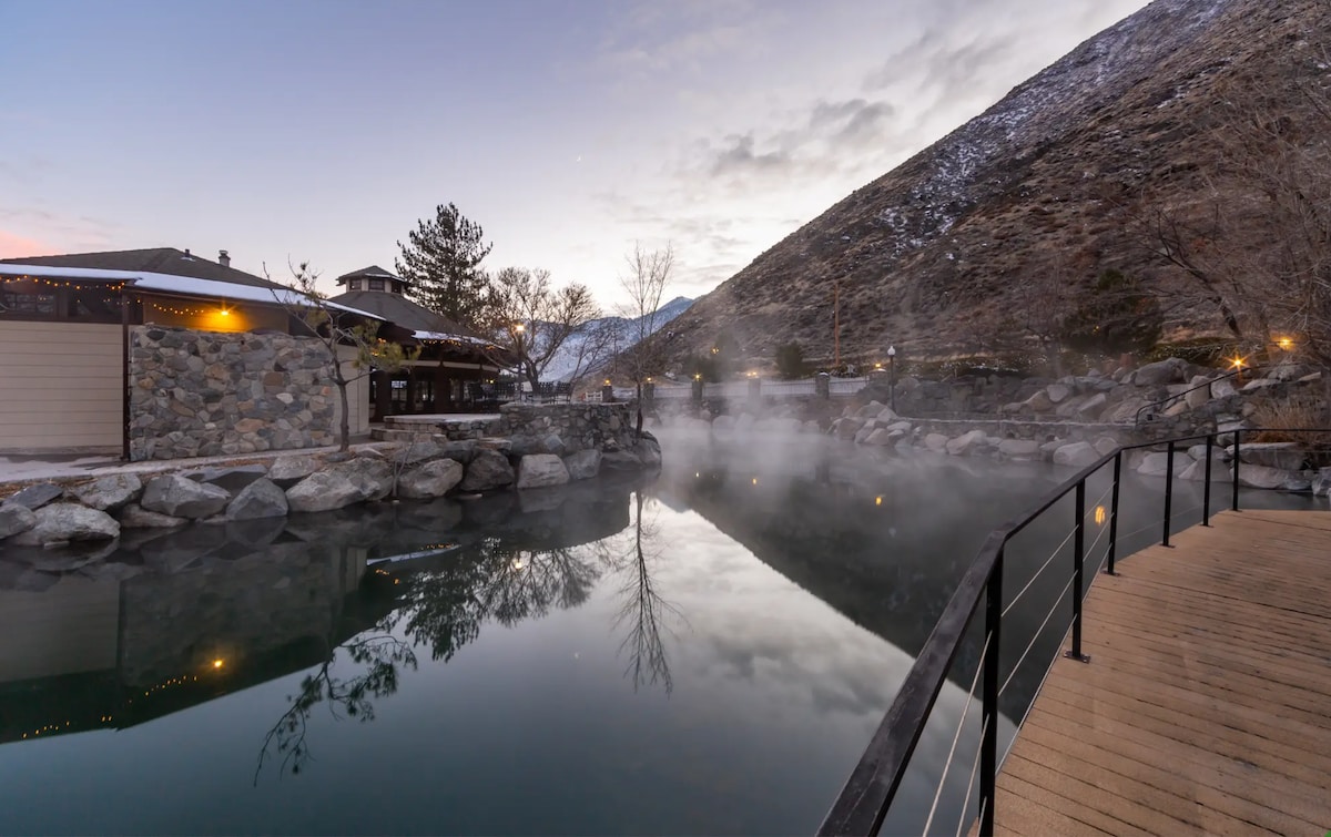 A serene landscape features a calm body of water surrounded by rocky formations. Steam rises gently from the surface, reflecting the soft hues of dusk. A wooden boardwalk extends into the scene, providing access to the tranquil setting and connecting to a nearby building illuminated by warm lights.
