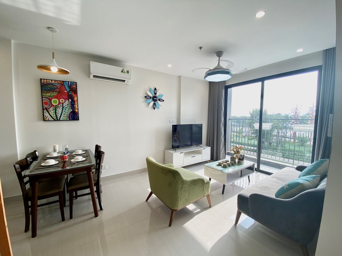 A modern living area includes a cozy green chair and a grey sofa, complemented by a dining table set for four. A large TV is positioned against the wall, while sliding glass doors provide access to a balcony with views of the outdoors. Decorative wall art adds a touch of color.