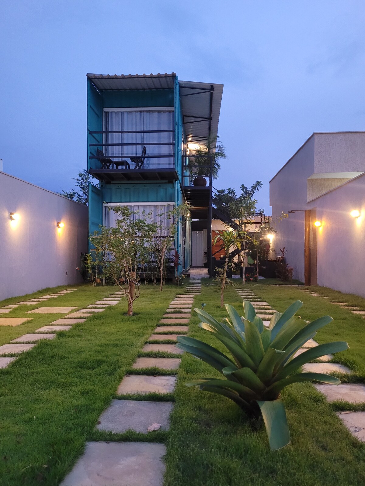 A two-story container loft is framed by landscaped greenery, featuring a pathway of stone tiles leading towards the entrance. Soft outdoor lighting illuminates the surrounding area, enhancing the peaceful ambiance. The patio area on the upper level includes seating that overlooks the garden.