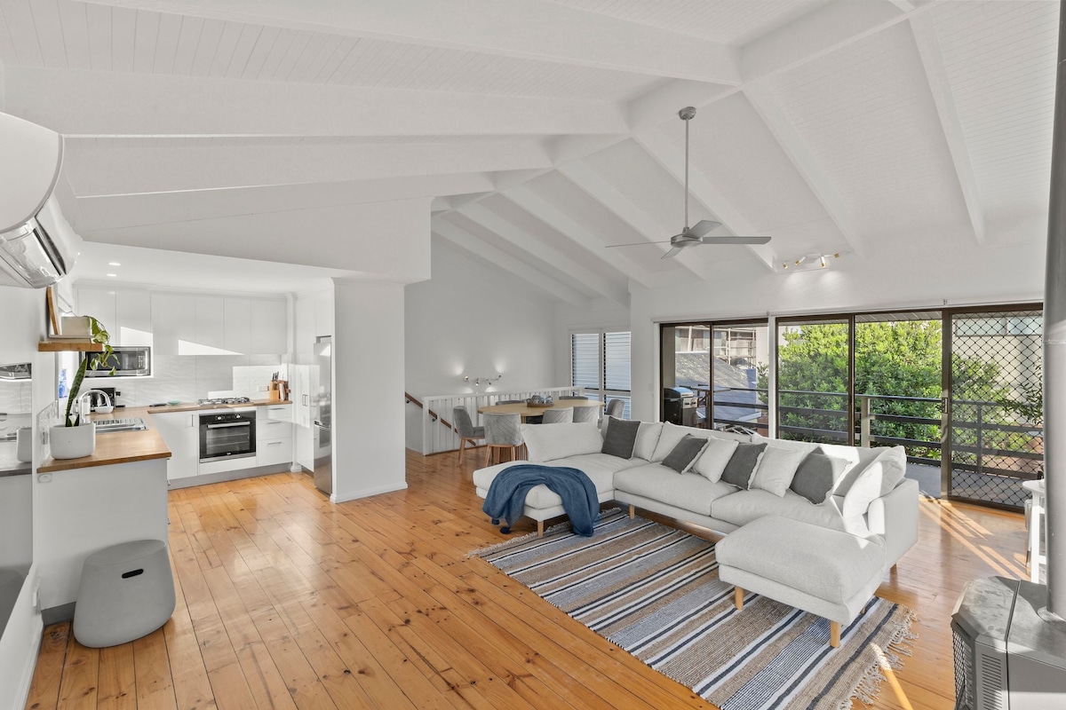 An open-plan kitchen and living area features timber floorboards and high ceilings with beams. A spacious modular couch is positioned on a striped area rug, while a modern kitchen with white cabinetry and stainless-steel appliances is visible in the background. Large windows provide natural light and views of the outside.