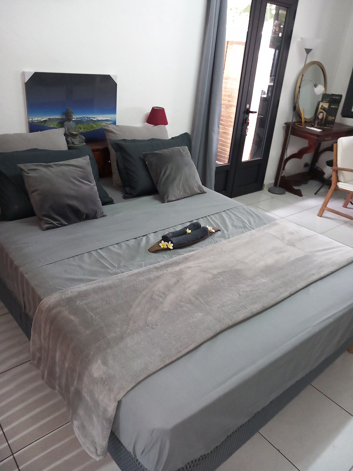 A queen-size bed is dressed in soft grey linens, accented with decorative pillows. The space features a bedside table with a lamp and an ornate mirror. Natural light enters through double doors, leading to a private outdoor area.