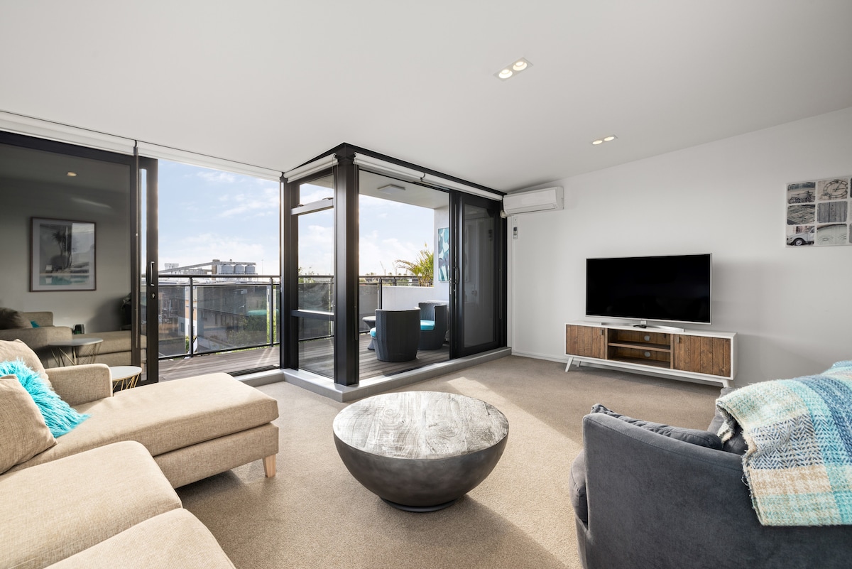A bright lounge area is visible, featuring a spacious sectional sofa and a contemporary coffee table. Large glass sliding doors allow natural light to fill the space and provide access to the outdoor deck. A smart TV is mounted on the wall opposite the seating.