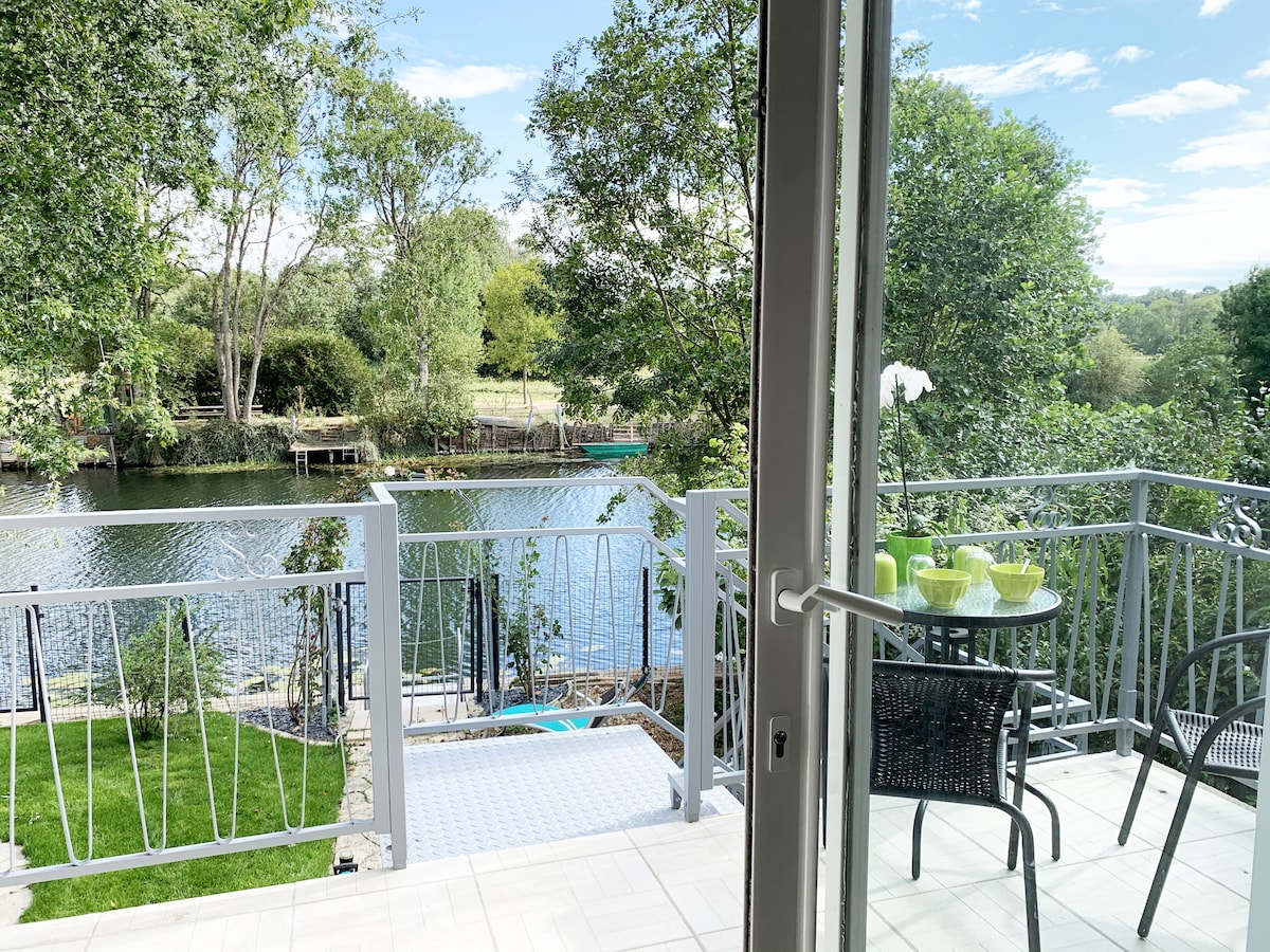 A view from the balcony showcases a serene river bordered by lush greenery. A small table with two chairs is placed on the terrace, while decorative dishes are set atop. The well-maintained garden is visible, along with a pathway leading to the water.
