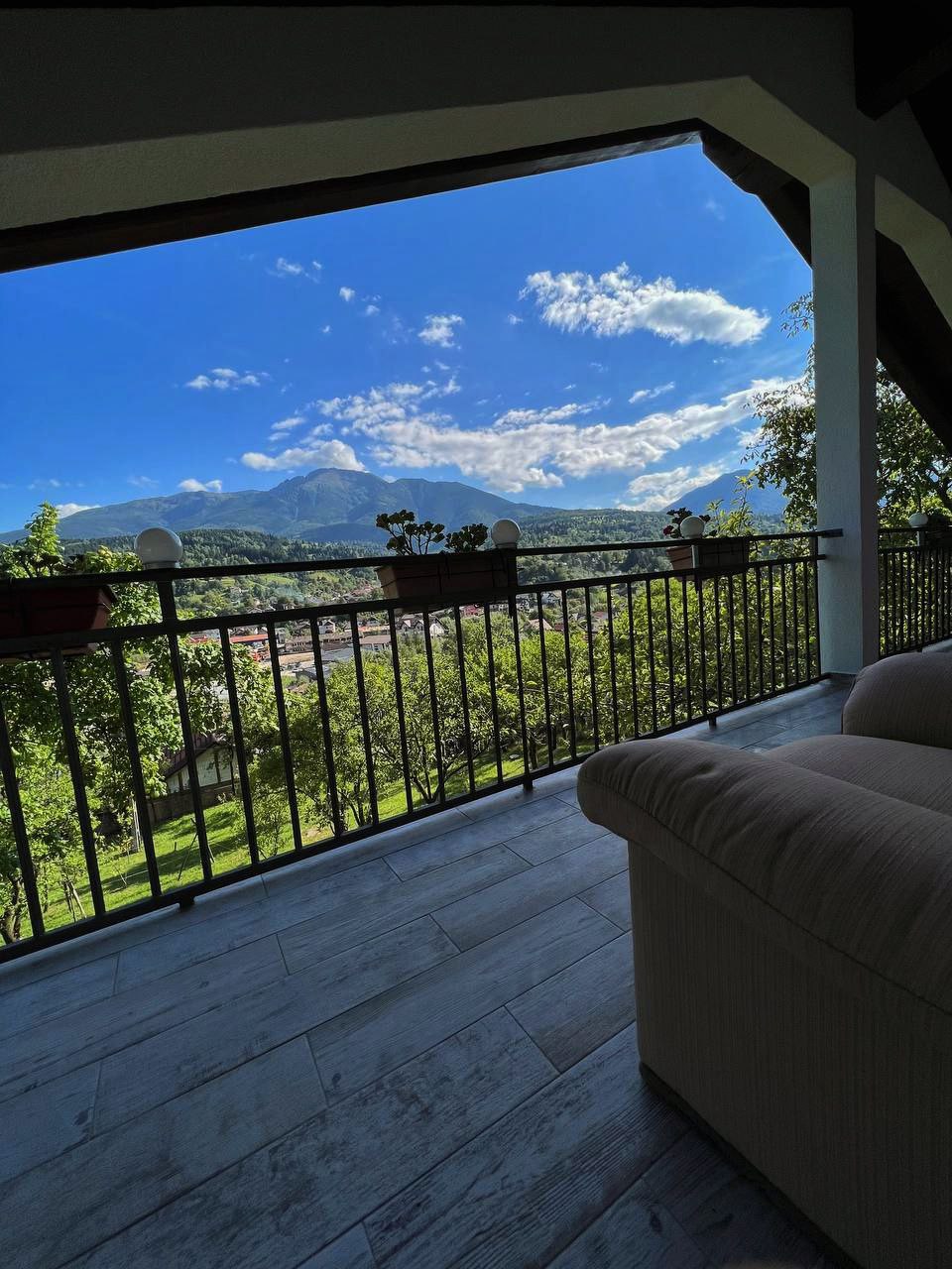 A covered balcony is shown, offering a scenic view of mountains and bright blue skies. Potted plants are positioned along the railing, and a light-colored sofa is visible, suggesting a comfortable outdoor seating area.