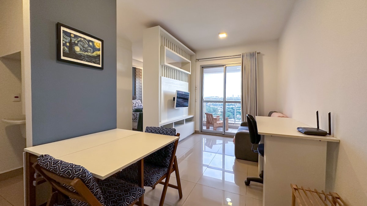 A modern living area features a large window offering expansive views. A small dining table with patterned chairs is positioned near the window. A desk and a chair are visible, along with a television mounted on a stylish shelf. Soft lighting provides a welcoming ambiance.