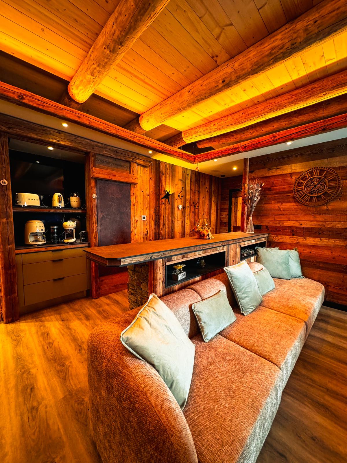 A cozy lounge area features a plush sofa adorned with soft, light-colored cushions. The room showcases wooden beams and paneling, creating a warm atmosphere. A bar-style counter is seen in the background, complemented by kitchen appliances, contributing to a well-equipped communal space.
