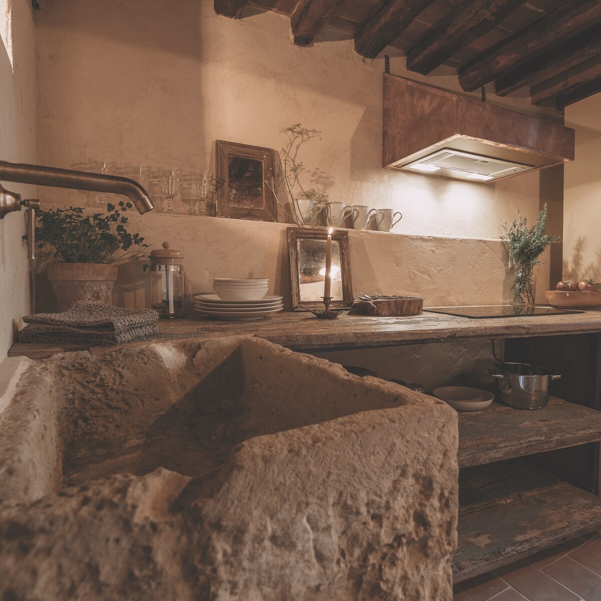 A rustic kitchen features stone countertops and an antique sink, blending history with functionality. Shelves hold dishware and glassware, while a candle adds a warm glow. Exposed wooden beams are visible overhead, enhancing the space's traditional charm.