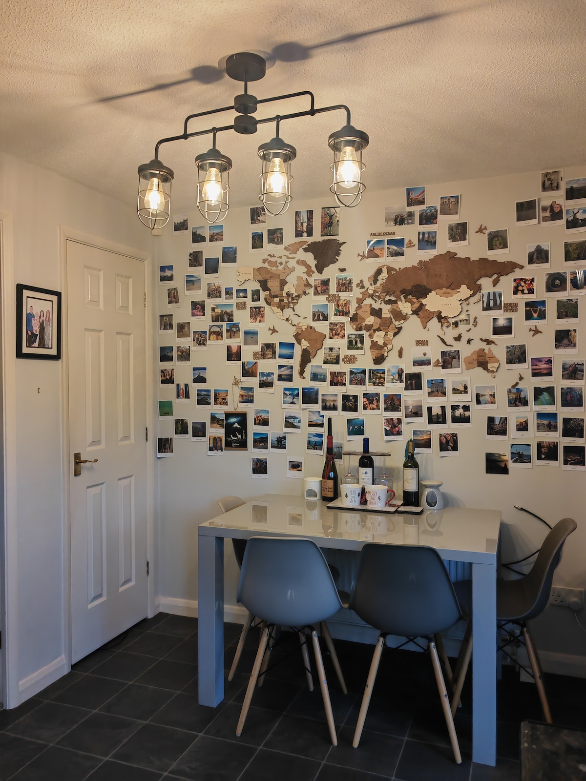 A dining area features a light-colored table surrounded by four modern chairs. A unique map made of cork outlines the world on the wall, adorned with numerous photographs capturing travel memories. Soft lighting comes from a stylish five-bulb fixture overhead.