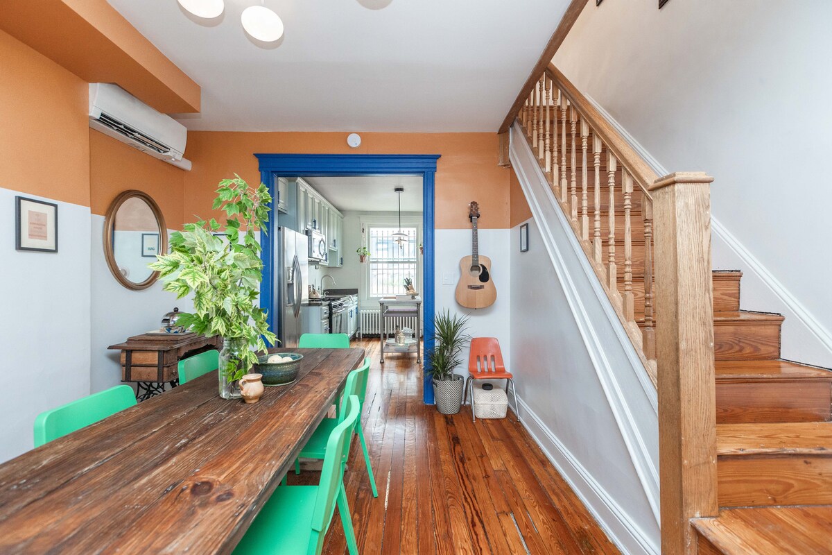 A bright dining area features a wooden table surrounded by green chairs, with light streaming in from nearby windows. The wall is painted in a warm orange, complementing the blue trim. A staircase leads to the upper level, and a glimpse of the kitchen is visible in the back.