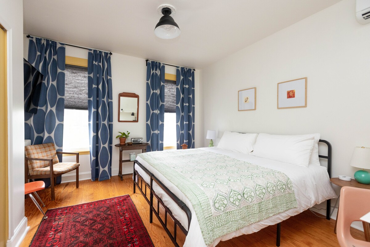 A bright bedroom features two spacious beds dressed in crisp white linens, accompanied by a decorative green throw. Light streams through stylish patterned curtains, while a small desk, a chair, and a mirror provide functional accents. A colorful area rug adds warmth to the hardwood floor.