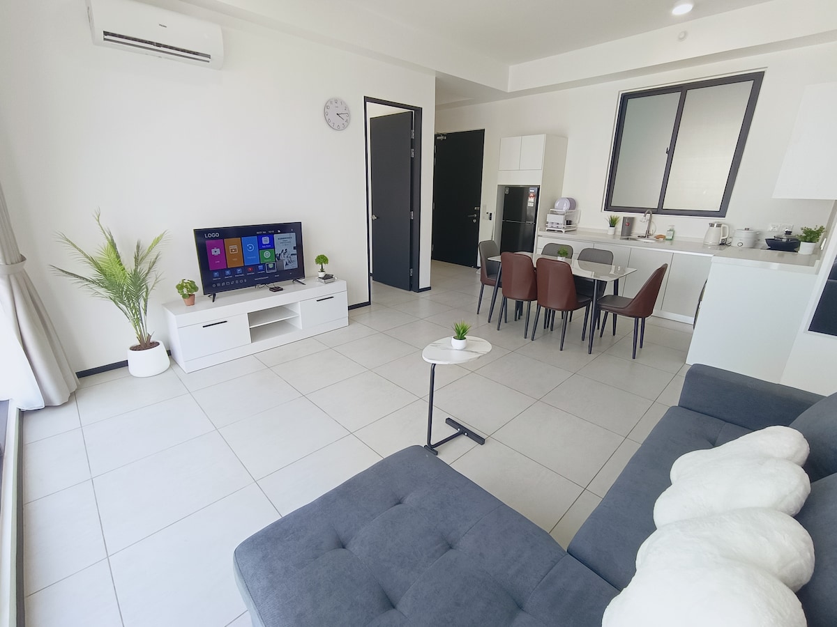 The living area showcases a spacious layout featuring a grey sectional sofa, a modern white TV stand with a television, and a dining table with six chairs. Natural light illuminates the space through large windows, complemented by soft decorative plants.