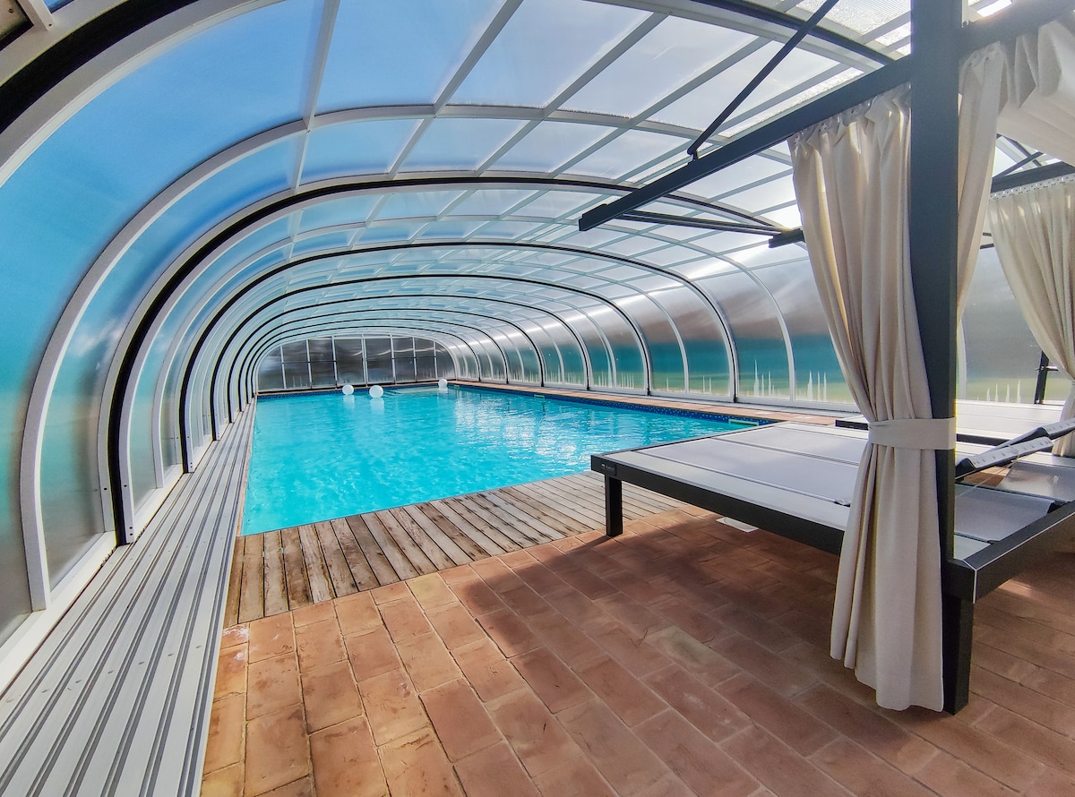 An indoor pool is enclosed by a curved transparent structure, allowing natural light to illuminate the space. Sun loungers are positioned alongside the pool, which reflects the clear blue water. Wooden decking surrounds the pool, creating a warm and inviting environment.