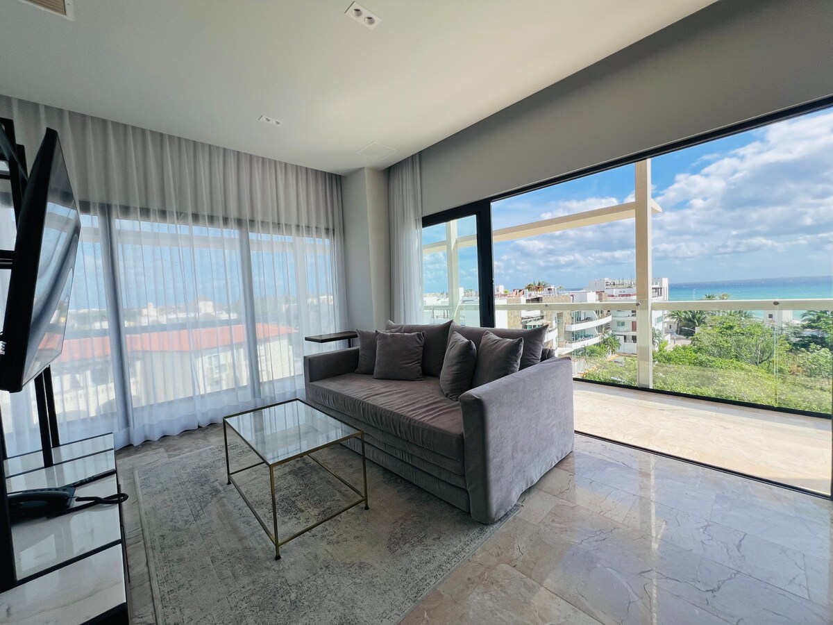 A spacious living area is highlighted by a large window, offering a view of the ocean and surrounding area. A comfortable gray sofa sits facing a glass coffee table, with sheer curtains allowing natural light to brighten the room.