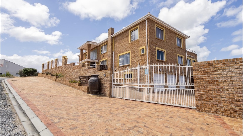 A double-storey home is presented with a sturdy brick exterior and large windows, set against a backdrop of blue sky and intermittent clouds. A gated entrance is visible, leading to a paved pathway, and a large decorative pot is positioned alongside the building.