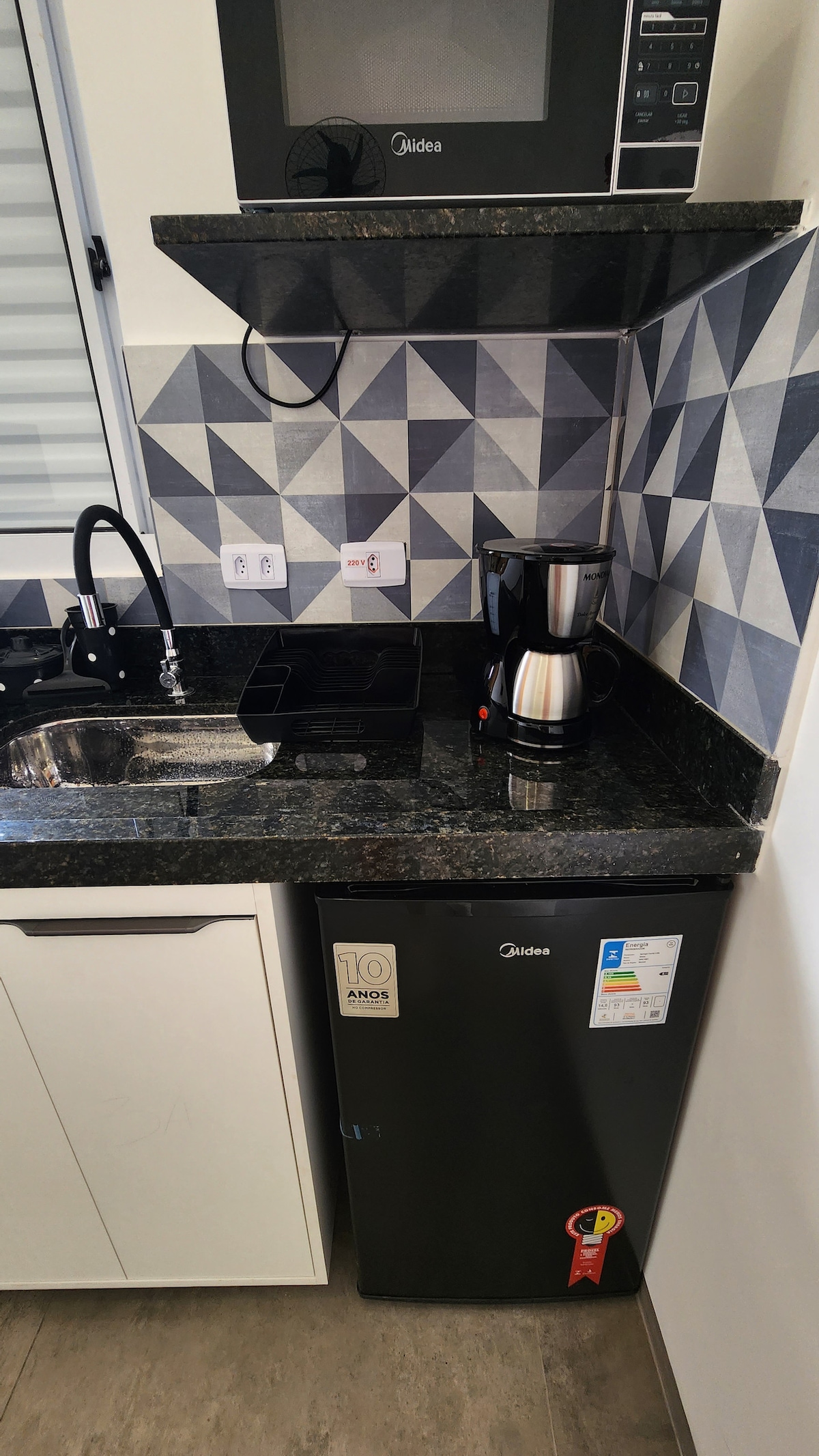 The kitchen area features a sleek black refrigerator and a coffee maker on a granite countertop. A microwave is mounted above the countertop, and geometric patterns adorn the wall, enhancing the modern look of the space.