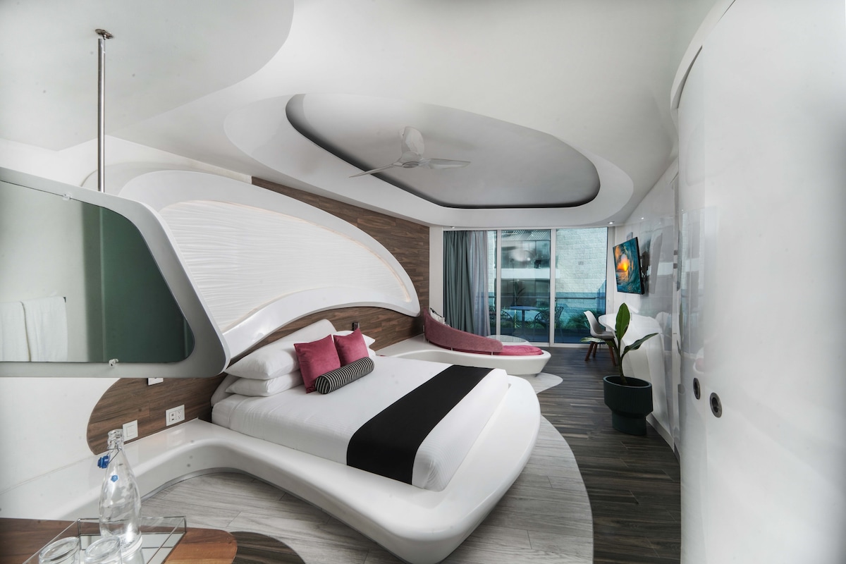 A contemporary suite design is featured, characterized by smooth, curved lines and a spacious bed with contrasting black and white bedding. A cozy seating area and large windows invite natural light, while a potted plant adds a touch of greenery to the space.