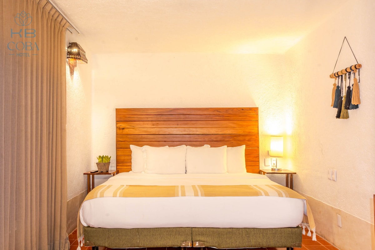 A spacious bed is positioned against a wooden headboard, complemented by neutral-toned bedding. Warm light from bedside lamps creates a restful ambiance. Soft curtains frame a window, and decorative elements, such as wall hangings, add a subtle touch of style to the room.