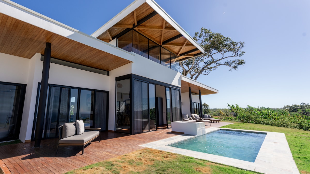 A modern villa is nestled on a hillside, showcasing large glass doors that create an open connection to the outdoors. A private pool is visible, surrounded by wooden decking, with lush greenery framing the view. The architectural design features a slanted roof and spacious balconies.