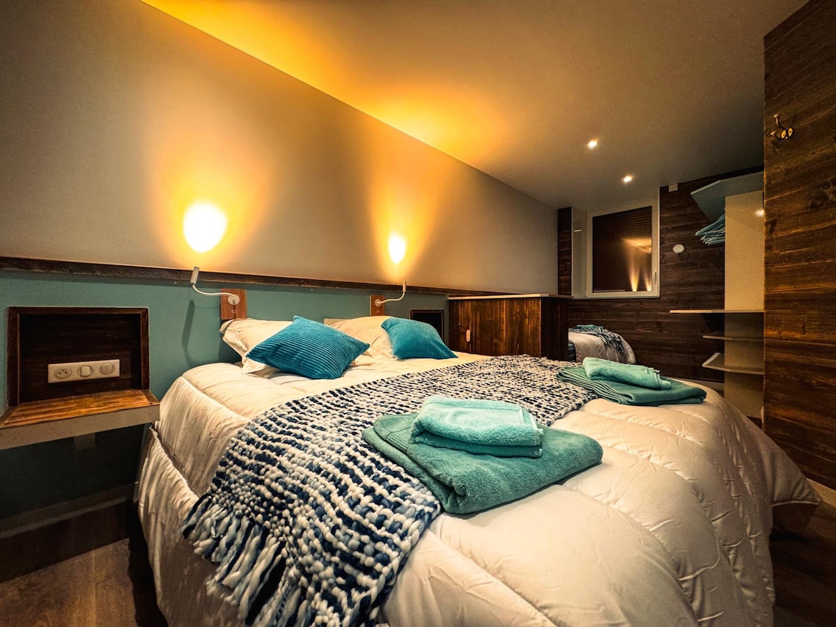 A cozy sleeping area is presented with a spacious bed adorned in neutral tones and accented by vibrant teal pillows. Soft lighting from wall fixtures creates a warm ambiance, complementing the wood features and shelving in the background.