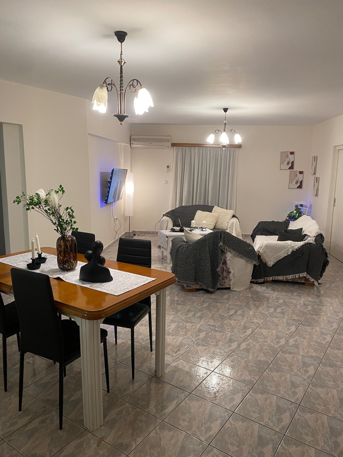 The living area features a dining table with four chairs and a cozy seating arrangement with two sofas covered in light-colored throws. A flat-screen TV is mounted on the wall, with soft ambient lighting enhancing the space. Large windows offer natural light.