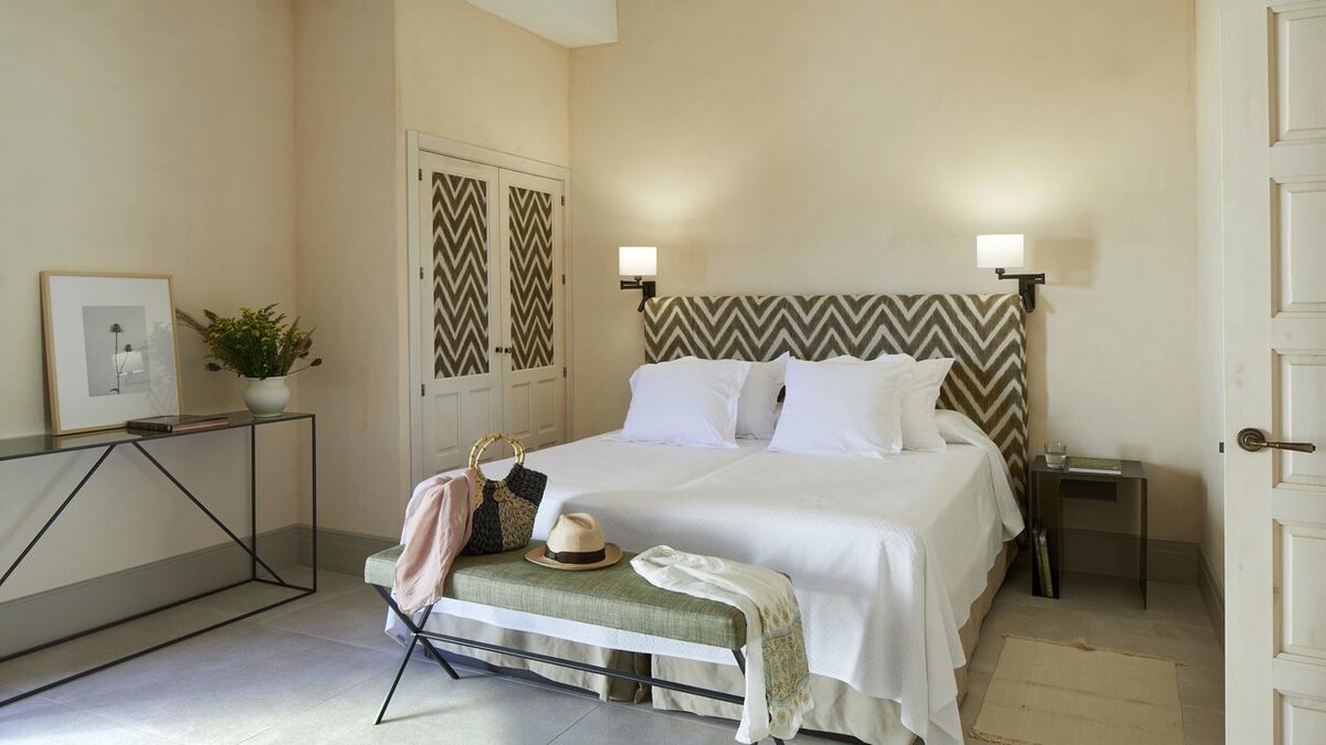 A bright bedroom features a spacious bed with white linens and two bedside lamps mounted on the walls. A bench sits at the foot of the bed, accompanied by a hat and a bag. A minimalist table and a large closet are also visible.