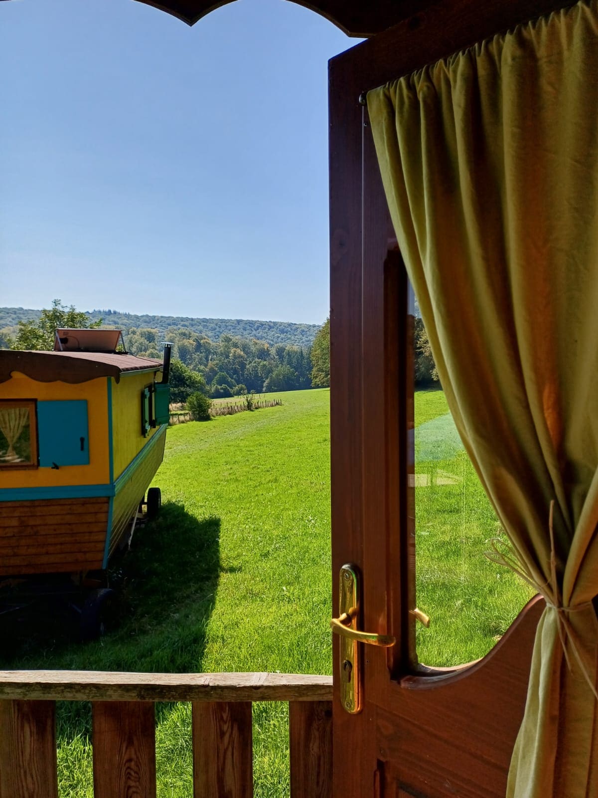 A doorway opens to a scenic view of vibrant green fields and distant hills, framed by a wooden entrance. Natural light bathes the space, and a colorful wagon is visible on the property, enhancing the rural charm.
