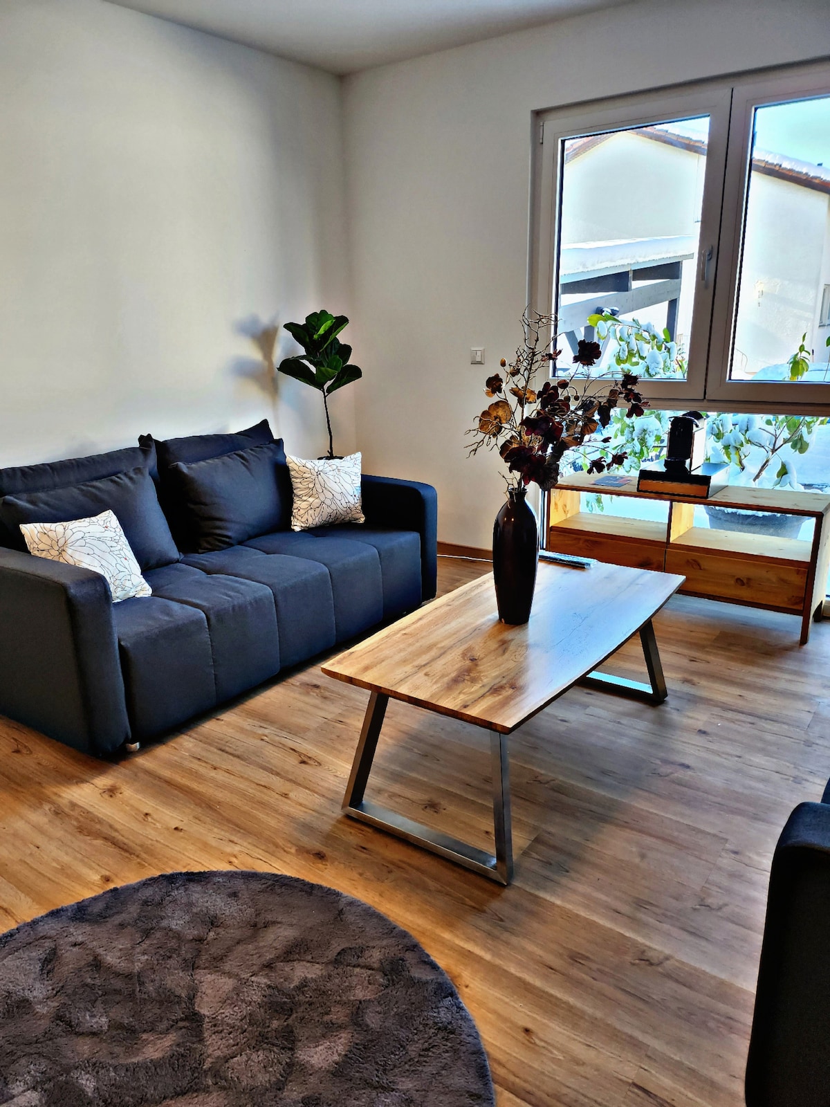 A living area features a dark sofa with decorative cushions, accompanied by a wooden coffee table. A floral arrangement in a black vase centers the table. Natural light enters through a large window, illuminating the space, while a potted plant enhances the decor.