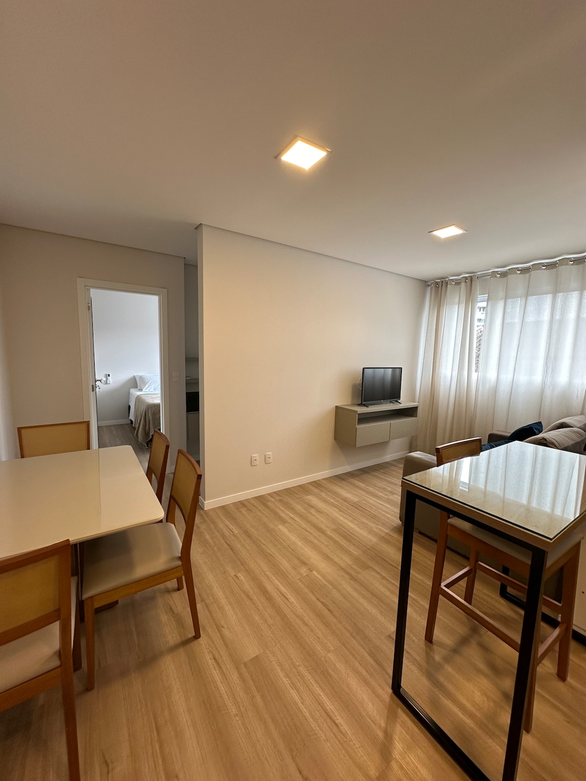 A modern living space is shown, featuring a smart TV mounted on the wall opposite a comfortable sofa. A light wooden dining table with several chairs is positioned to the left. Natural light filters through sheer curtains, creating a bright and inviting atmosphere.