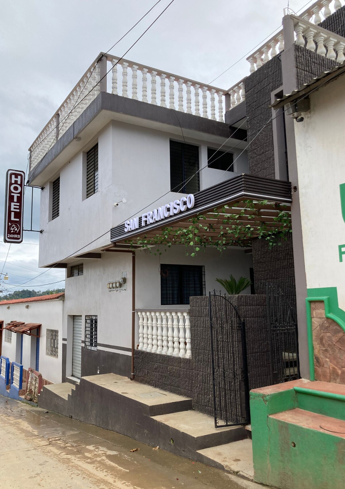 A two-story building labeled 'Hotel San Francisco' is depicted, featuring a modern facade with white walls and decorative balconies. Greenery is visible near the entrance, which is framed by a wrought-iron gate. The street setting includes neighboring structures and an unobstructed view of the hotel.