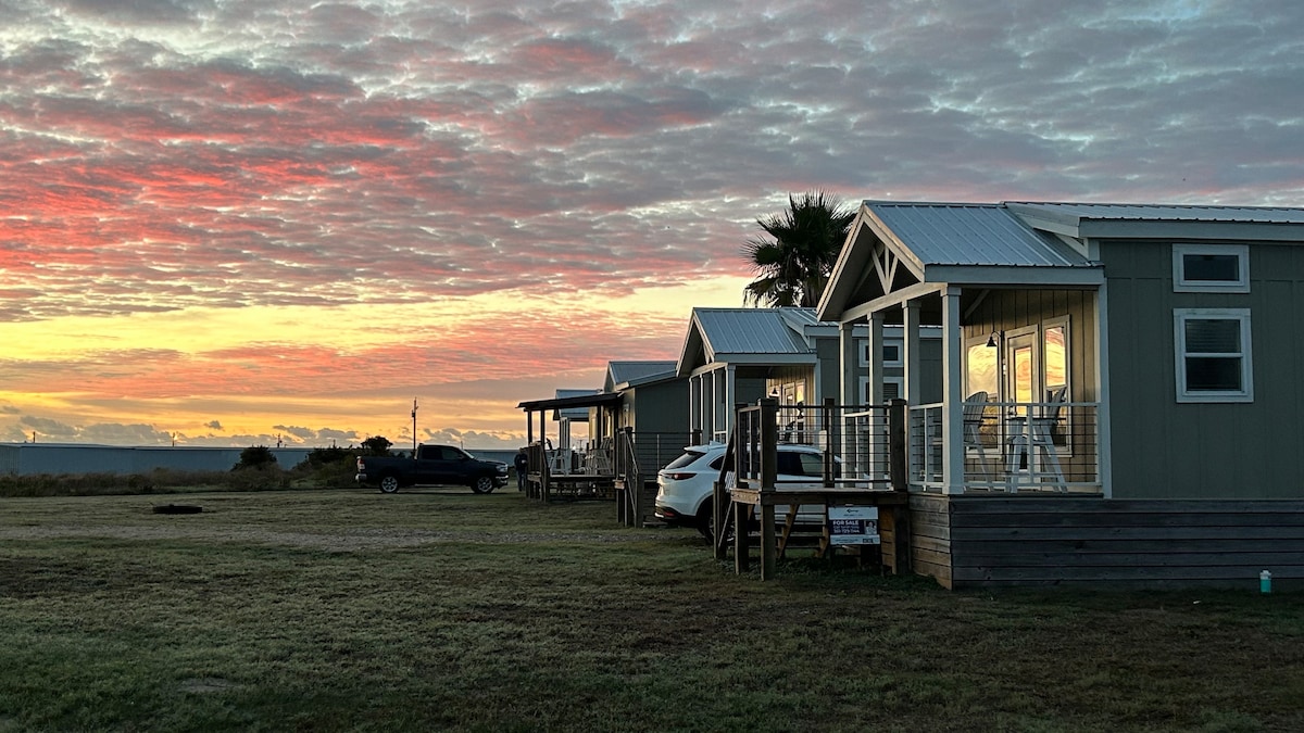 The cabin units are positioned side by side, featuring large front porches with glass doors. A vibrant sunset casts warm colors over the scene, creating a serene backdrop. A few vehicles are visible in the foreground, with expansive grassy areas surrounding the cabins.