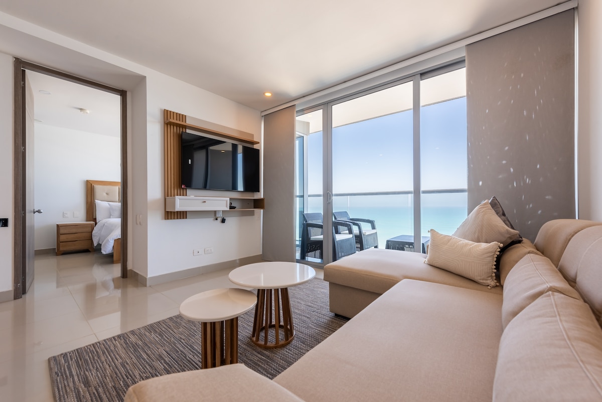 A modern living area is featured, equipped with a spacious sectional sofa and two round coffee tables. Large glass doors provide sweeping views of the beach and ocean. Soft natural light fills the space, creating a calming atmosphere. A bedroom is visible in the background.