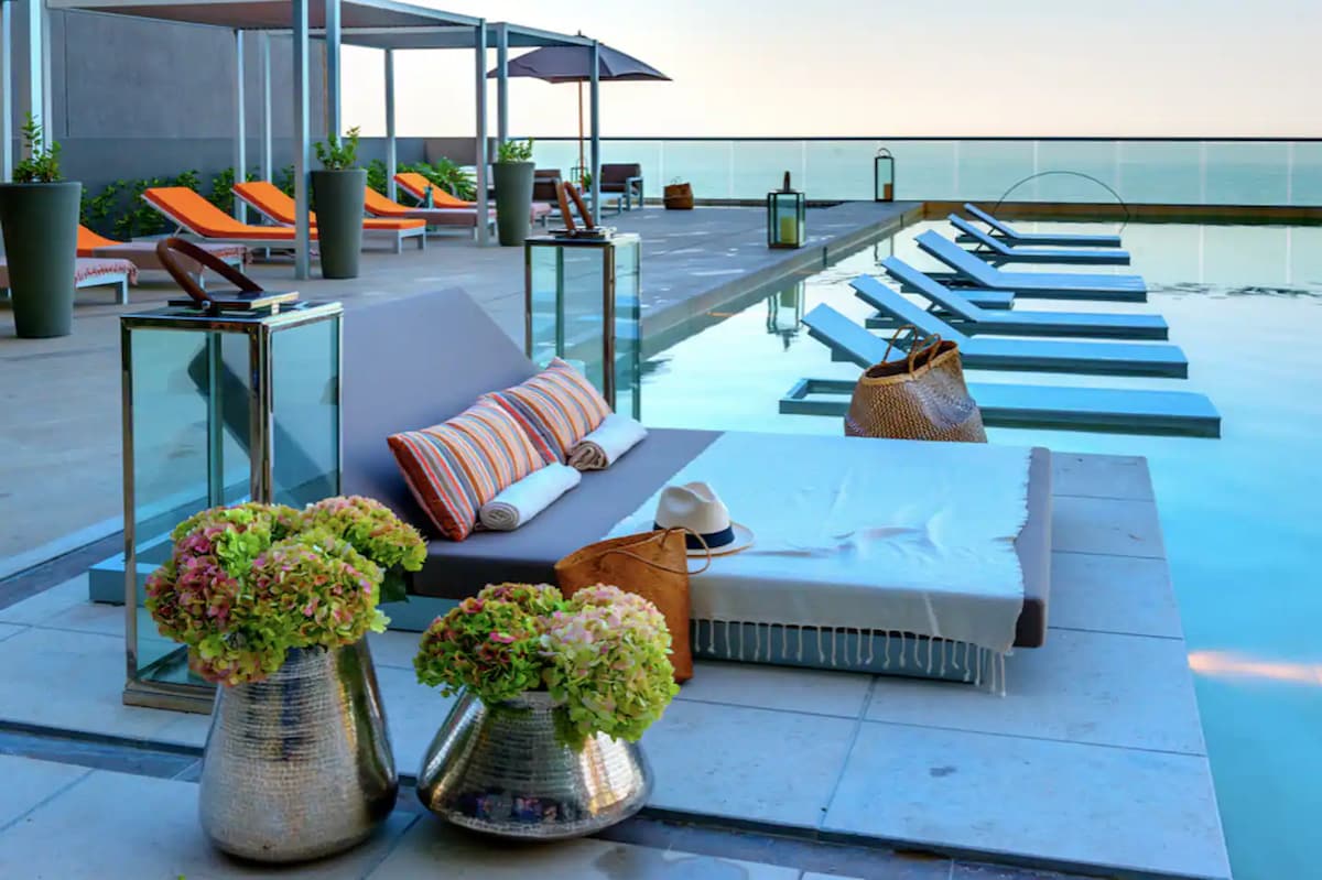 A modern pool area features comfortable lounge chairs arranged alongside the water, with shades of orange and gray providing a vibrant touch. Two stylish planters filled with lush flowers add a natural element, while soft towels and a hat rest on a sunbed, inviting relaxation.