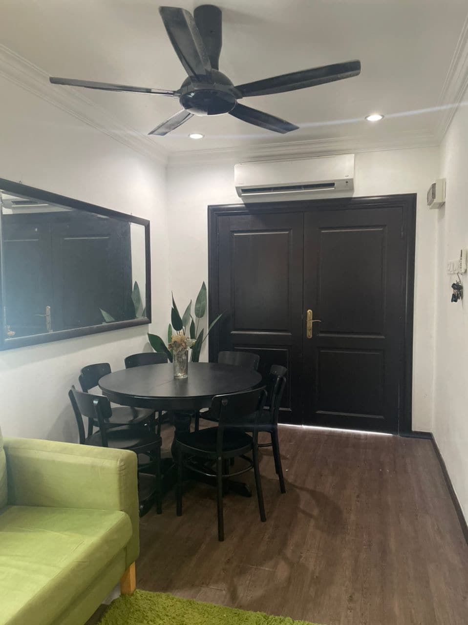 A dining area features a round black table surrounded by four chairs. A green sofa is positioned to the side, with a textured rug beneath. A large mirror reflects the space, while potted plants add a touch of greenery. Ceiling fan and wall-mounted air conditioning unit are visible.