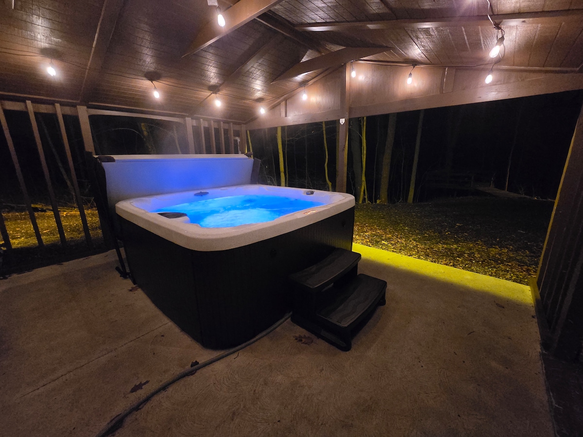 An inviting hot tub is located under a covered structure, illuminated by soft overhead lights. The water glows in shades of blue, creating a relaxing ambiance. Surrounding trees are lightly visible, contributing to the secluded atmosphere.