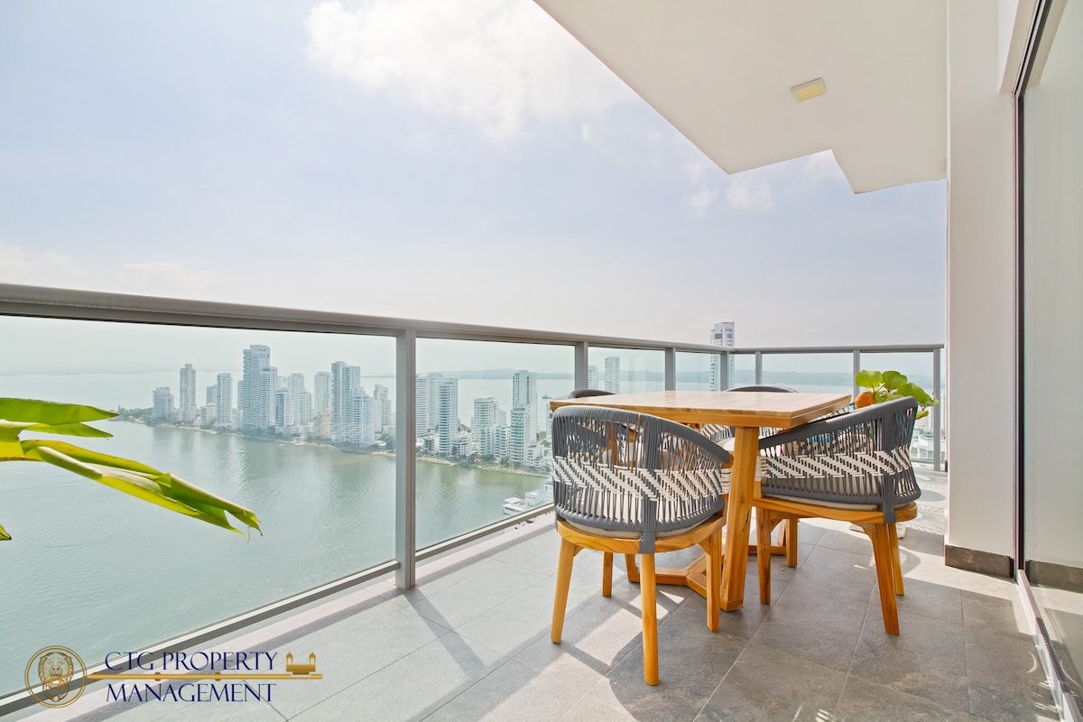 An outdoor dining area is presented on the spacious balcony, featuring a wooden table accompanied by stylish chairs. A panoramic view of the bay and city skyline is visible beyond the railing, with a clear sky enhancing the tranquil ambiance.