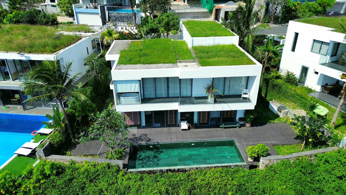 The villa is surrounded by lush greenery, featuring a rooftop garden. An inviting infinity pool lies adjacent to the structure, while the outdoor decking provides spaces for relaxation. Large glass windows reflect natural light, enhancing the contemporary architectural design.