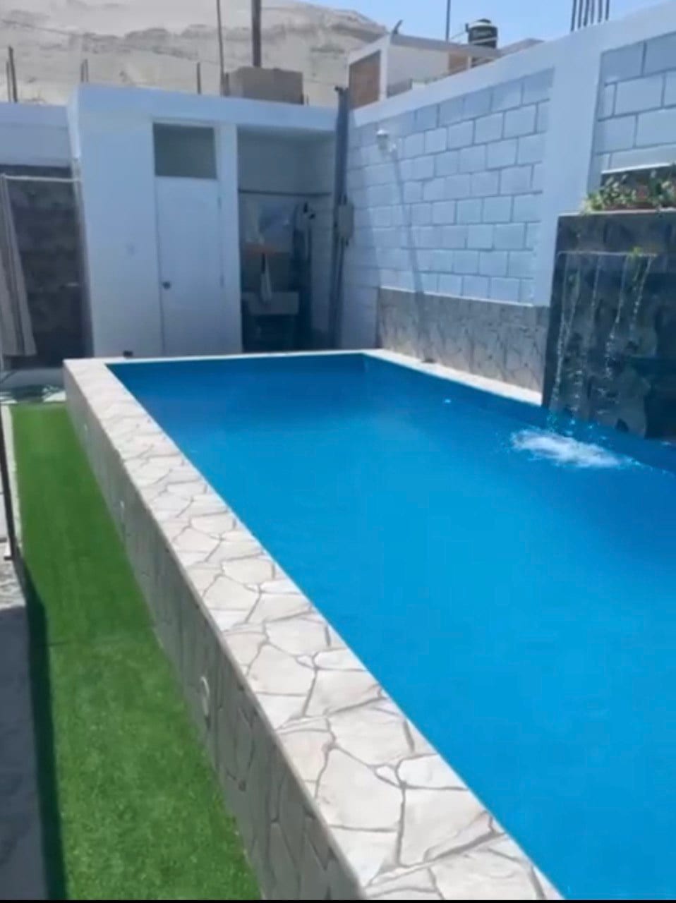 A rectangular swimming pool features crystal-clear water and a gentle waterfall cascading down a stone wall. The surrounding area includes textured stonework and artificial green grass, providing a refreshing ambiance. A door leads to an enclosed space nearby, emphasizing the pool's serene setting.