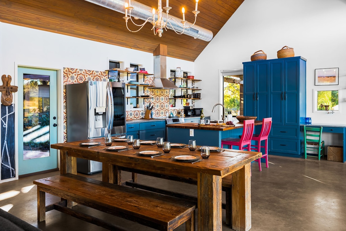 An open-concept kitchen showcases a large wooden dining table, seating ten, surrounded by vibrant chairs. The kitchen features blue cabinetry, modern appliances, and decorative tiles. Natural light filters through multiple windows, illuminating the inviting space.