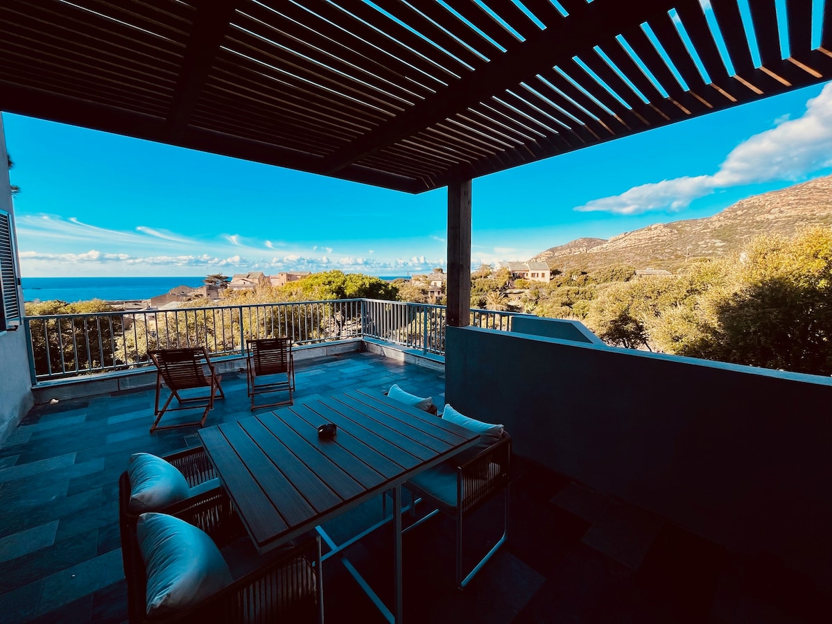 A spacious terrace presents views of the sea and mountains, framed by wooden beams above. A wooden dining table is accompanied by two chairs, creating an inviting space for relaxation. Lush greenery is visible in the background, enhancing the serene atmosphere.