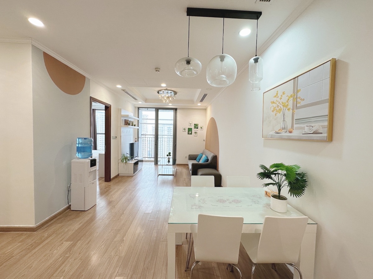 An open living space features a modern dining table surrounded by white chairs. A comfortable sofa is positioned near a large window, allowing natural light to fill the area. Decorative elements include a potted plant and wall art, enhancing the inviting ambiance.