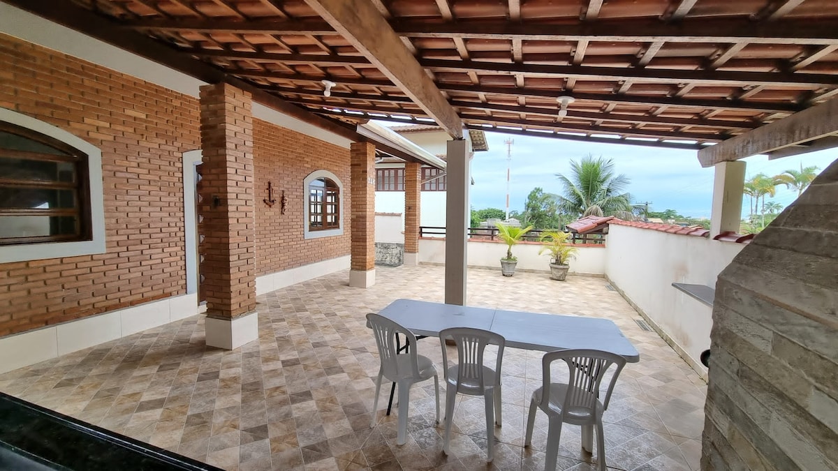 A spacious outdoor patio features tiled flooring with a wooden ceiling overhead. A rectangular table with three white chairs is positioned in the center, surrounded by potted plants. The area opens to views of greenery and the sky, offering a relaxed setting for gatherings.