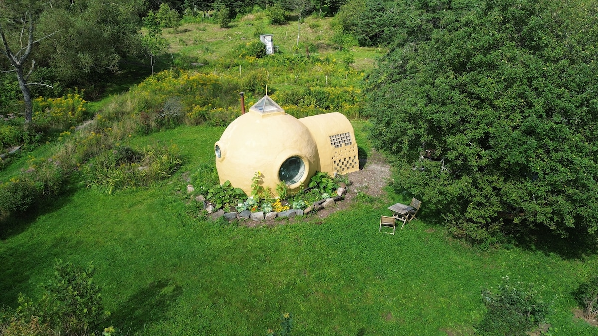 A spherical dome structure is surrounded by lush greenery and vibrant wildflowers. Natural stone landscaping highlights the perimeter, while a small chair sits nearby, offering a space for relaxation. The dome features a large window and a chimney, blending with the tranquil outdoor setting.