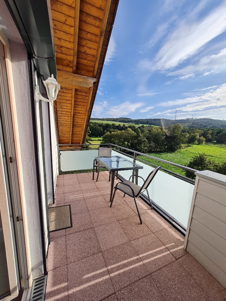 A private balcony overlooks lush green fields and rolling hills. A glass-top table is accompanied by a single chair, and the sloped wooden ceiling adds an architectural detail. Natural light illuminates the space, enhancing the serene outdoor setting.