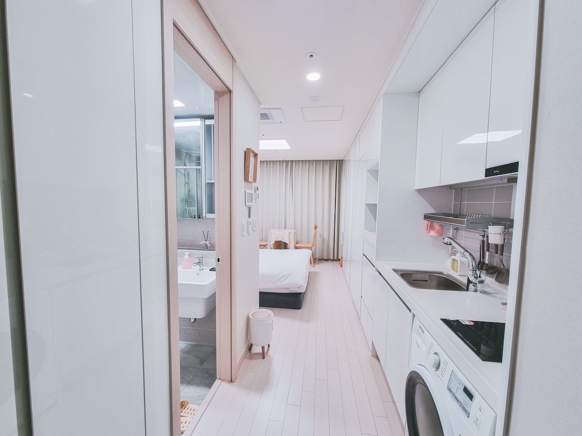 A modern, well-lit hallway leads to a cozy sleeping area featuring a queen size bed and soft bedding. An equipped kitchenette and laundry facilities are visible, with sleek cabinetry and kitchen appliances enhancing functionality.