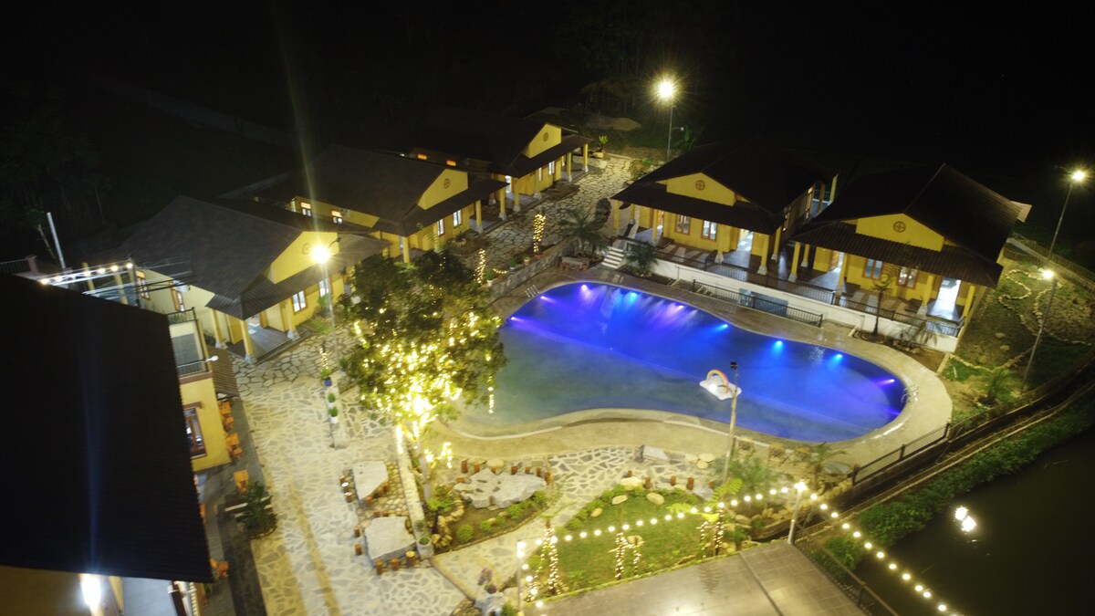 The image captures a night view of the property, featuring a spacious swimming pool illuminated in soft blue. Surrounding buildings with warm yellow accents provide accommodation, while garden pathways and seating areas enhance the outdoor ambiance. Soft lighting highlights the tranquil setting.
