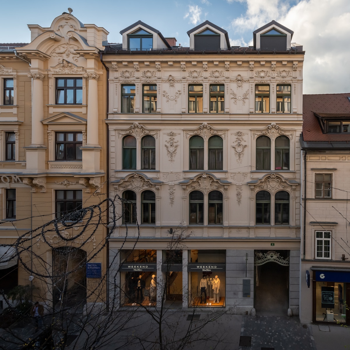 The image captures the exterior of a historic building featuring ornate architectural details and decorative elements. Large windows allow natural light to flood the interior. The structure is framed by neighboring buildings, highlighting its distinctive style and central location within the urban environment.