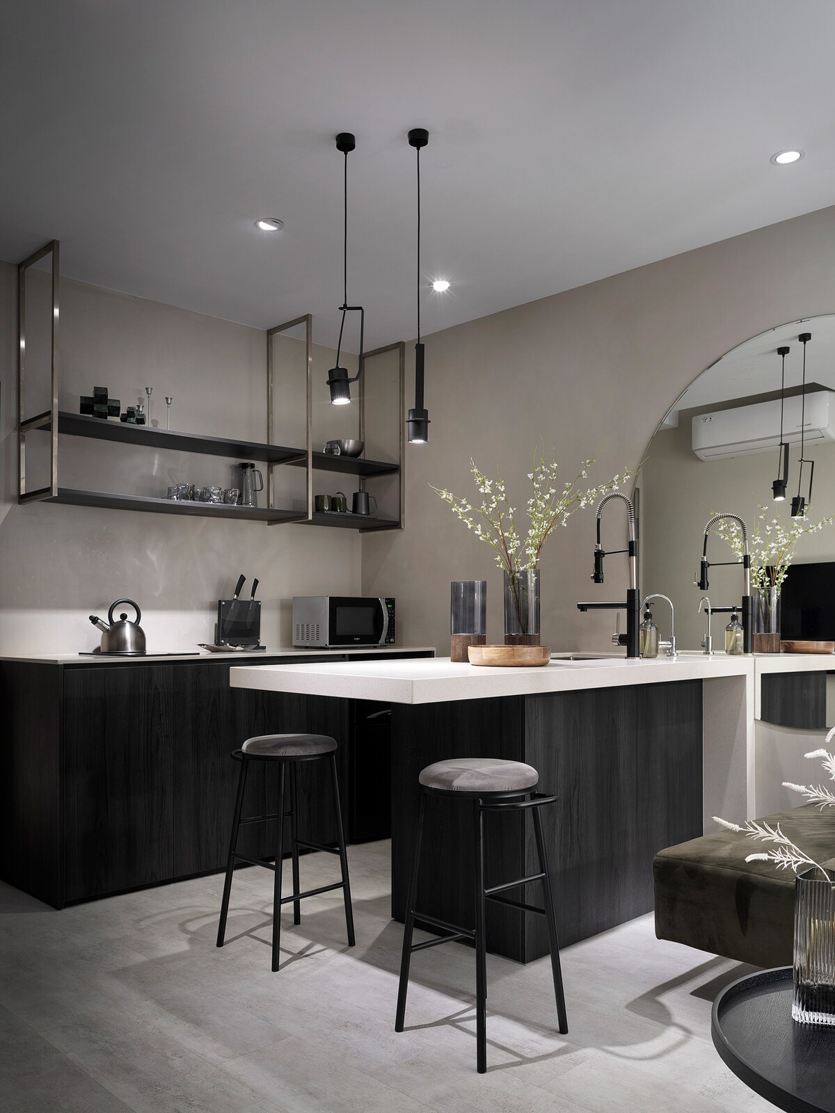A modern kitchen features dark cabinetry paired with light countertops. Open shelving displays glassware, while pendant lights hang above a compact dining area with two stools. A microwave and kettle are positioned on the counter, complemented by decorative floral arrangements.