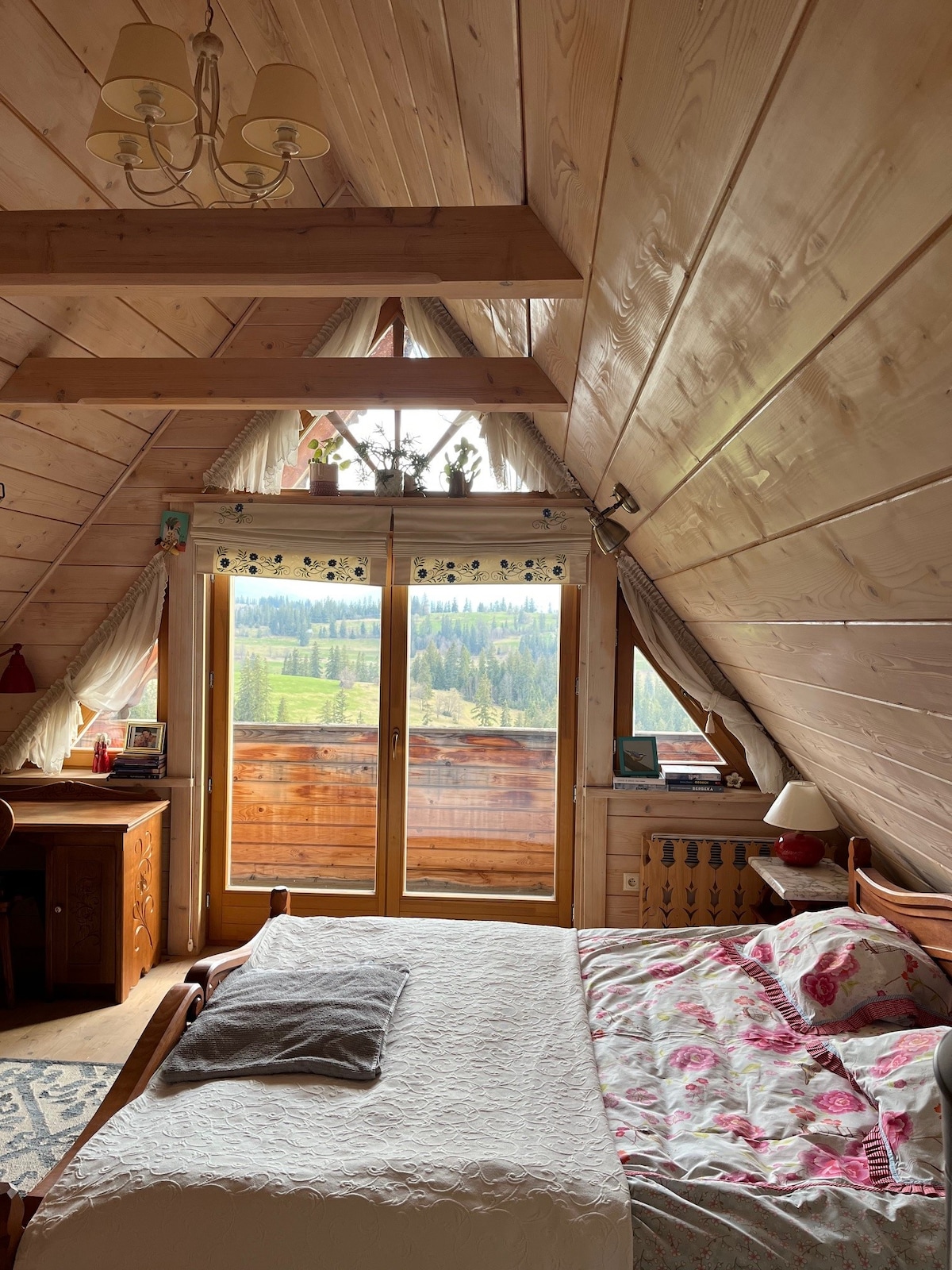 A wooden-paneled bedroom features a spacious bed dressed in floral bedding. Large glass doors lead to a balcony, offering views of the surrounding greenery. A writing desk sits beside the bed, and decorative curtains frame the windows, enhancing the room's cozy ambiance.