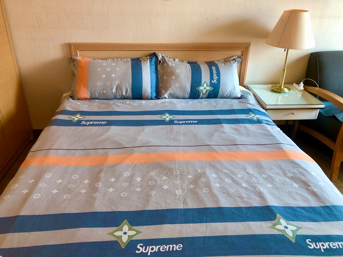 A spacious bed is dressed in a patterned gray and blue duvet cover. Two pillows display a repeating motif, while a bedside lamp provides soft lighting. A nearby side table is positioned for convenience, complementing the room's functional design.